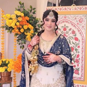 May include: A woman in a traditional South Asian outfit, featuring a white and gold embroidered tunic and matching pants. A navy blue patterned shawl is draped over the shoulders. The ensemble is accessorized with a headpiece and jewelry. A bouquet of yellow and orange flowers is in the background.