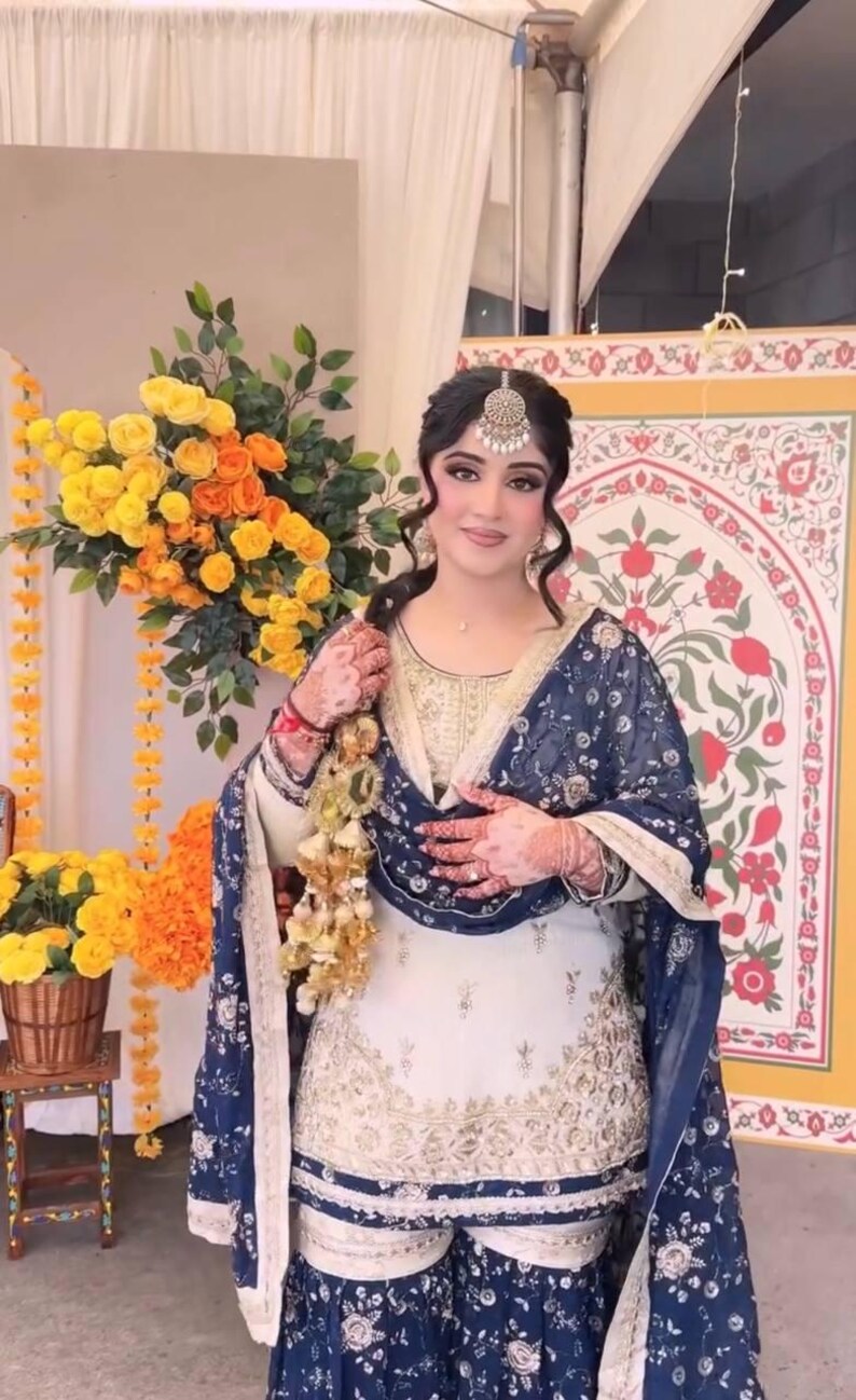 May include: A woman in a traditional South Asian outfit, featuring a white and gold embroidered tunic and matching pants. A navy blue patterned shawl is draped over the shoulders. The ensemble is accessorized with a headpiece and jewelry. A bouquet of yellow and orange flowers is in the background.