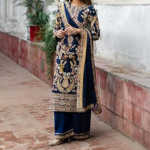 Può includere: Un completo formale ricamato blu navy. L'ensemble comprende una tunica a maniche lunghe, pantaloni a gamba larga e una sciarpa abbinata. L'outfit è impreziosito da ricami e finiture color oro e crema. L'outfit è indossato con scarpe color oro.