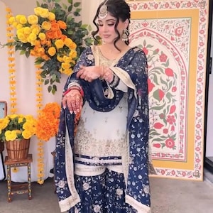 May include: A traditional South Asian outfit, featuring a white and navy blue floral print salwar kameez. The ensemble includes a matching dupatta and is adorned with gold embroidery. The outfit is worn with jewelry and henna.