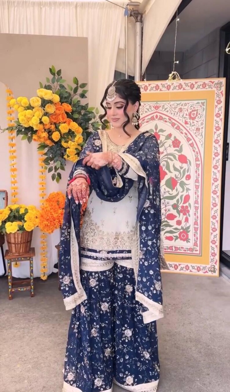 May include: A traditional South Asian outfit, featuring a white and navy blue floral print salwar kameez. The ensemble includes a matching dupatta and is adorned with gold embroidery. The outfit is worn with jewelry and henna.