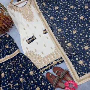 May include: An elegant white and gold embroidered salwar kameez set with a matching navy blue dupatta. The set includes a long tunic, wide-legged pants, and a scarf. Brown sandals with embellishments and pink flowers complete the ensemble.