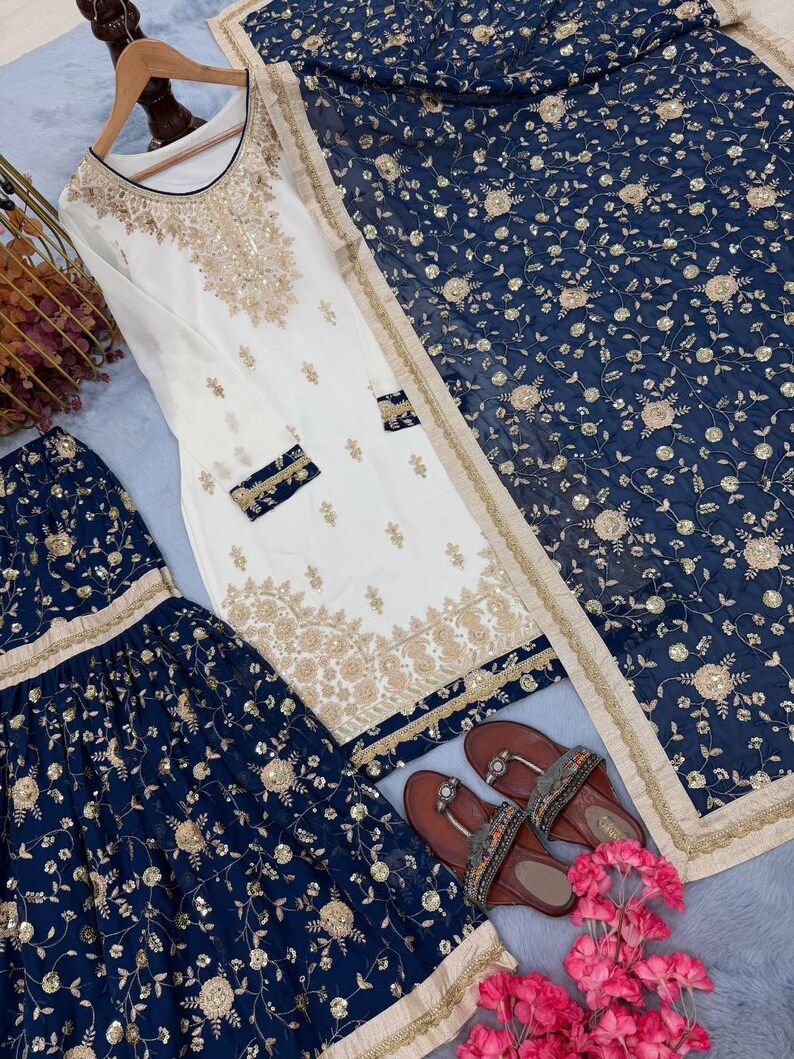 May include: An elegant white and gold embroidered salwar kameez set with a matching navy blue dupatta. The set includes a long tunic, wide-legged pants, and a scarf. Brown sandals with embellishments and pink flowers complete the ensemble.