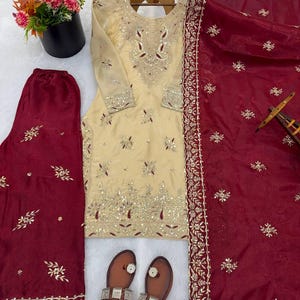 Puede incluir: Un conjunto salwar kameez bordado de color crema, con un dupatta y pantalones burdeos a juego. El conjunto incluye una t&uacute;nica de manga larga con intrincados adornos dorados y plateados. Tambi&eacute;n se incluyen sandalias de cuero marr&oacute;n con detalles plateados.