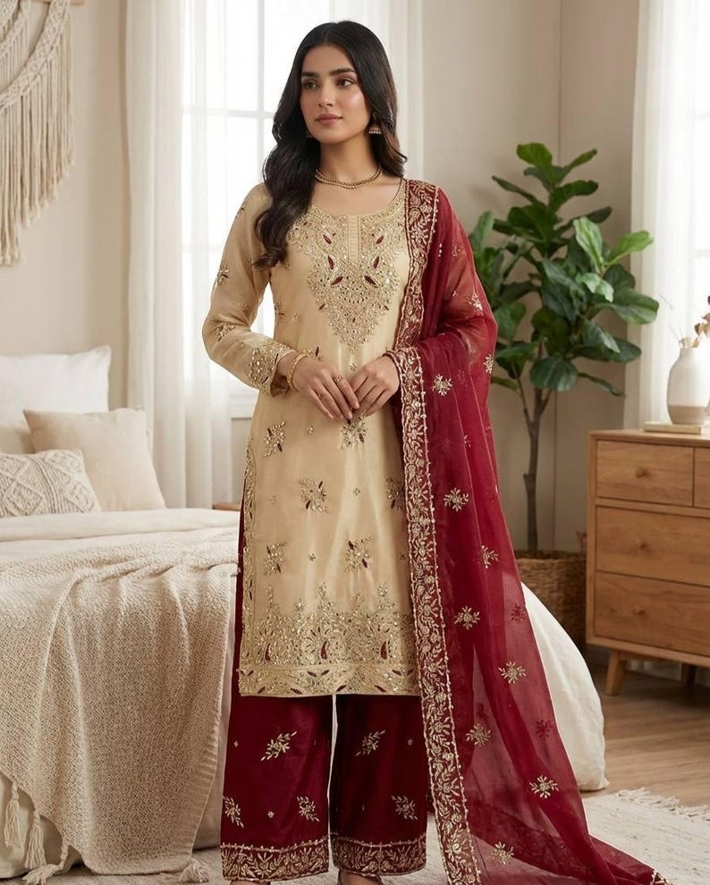 Puede incluir: Un conjunto salwar kameez beige bordado con un dupatta granate. La t&uacute;nica de manga larga y los pantalones de pernera ancha est&aacute;n adornados con bordados dorados. El dupatta granate presenta un borde bordado a juego.