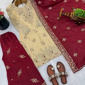 Puede incluir: Un conjunto salwar kameez bordado de color crema con una dupatta burdeos y pantalones a juego. El atuendo presenta intrincados adornos dorados y plateados. Sandalias marrones con detalles plateados completan el conjunto. El conjunto se muestra sobre una superficie blanca.