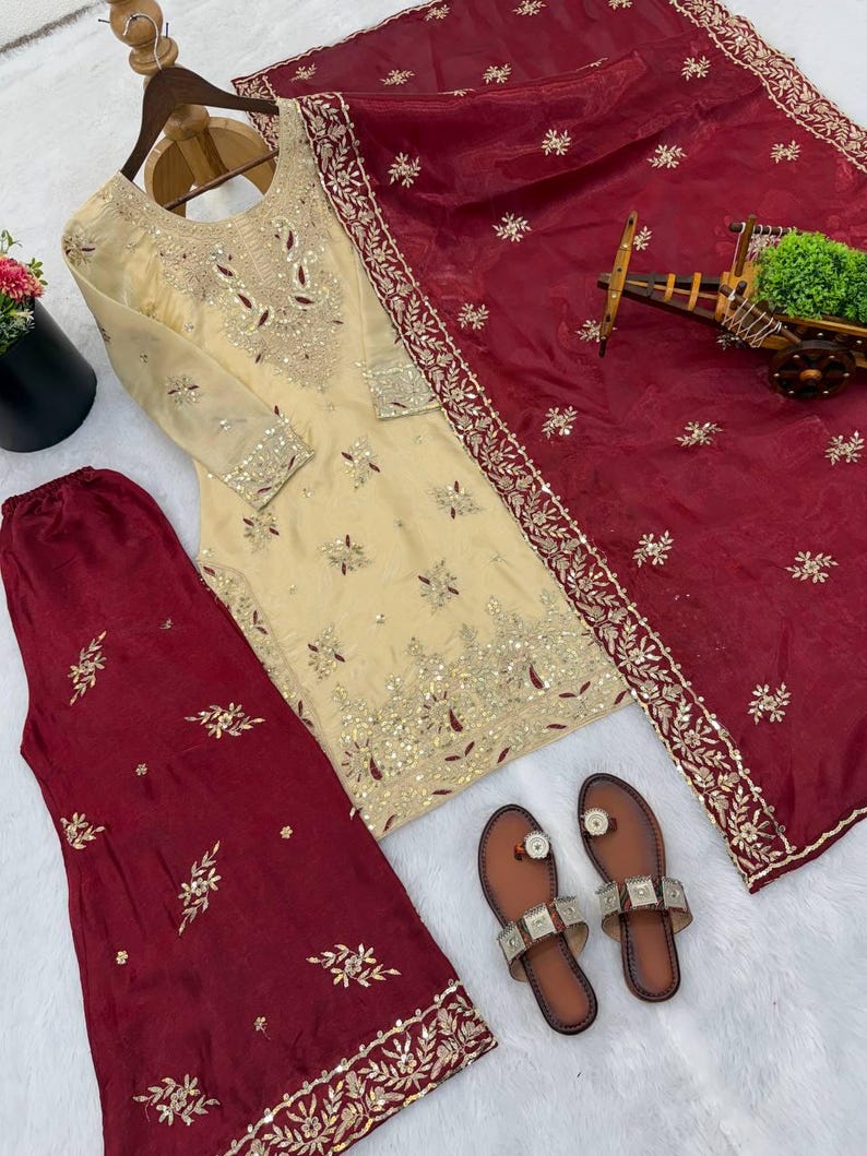 Puede incluir: Un conjunto salwar kameez bordado de color crema con una dupatta burdeos y pantalones a juego. El atuendo presenta intrincados adornos dorados y plateados. Sandalias marrones con detalles plateados completan el conjunto. El conjunto se muestra sobre una superficie blanca.