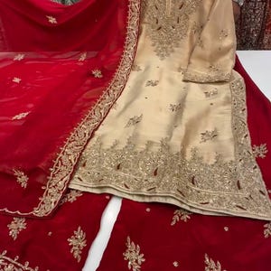 Puede incluir: Un conjunto salwar kameez bordado de color crema con una dupatta roja y pantalones a juego. El conjunto presenta intrincados bordados dorados en el escote, las mangas y el dobladillo. La dupatta roja tiene un borde bordado en oro. El atuendo se exhibe sobre una superficie blanca.