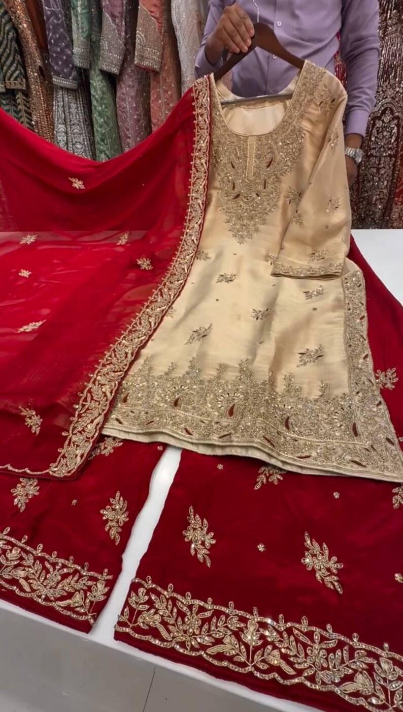 Puede incluir: Un conjunto salwar kameez bordado de color crema con una dupatta roja y pantalones a juego. El conjunto presenta intrincados bordados dorados en el escote, las mangas y el dobladillo. La dupatta roja tiene un borde bordado en oro. El atuendo se exhibe sobre una superficie blanca.