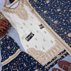 May include: An elegant white and navy blue embroidered salwar kameez set. The white tunic features intricate gold embroidery and embellishments. The navy blue pants and dupatta are adorned with floral patterns and gold accents. The set includes a pair of brown sandals.