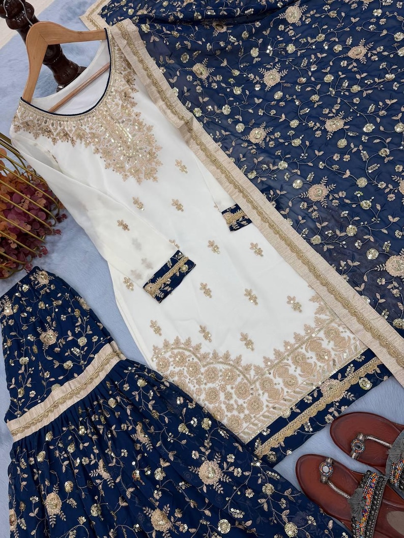 May include: An elegant white and navy blue embroidered salwar kameez set. The white tunic features intricate gold embroidery and embellishments. The navy blue pants and dupatta are adorned with floral patterns and gold accents. The set includes a pair of brown sandals.
