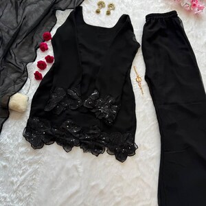 May include: A black two-piece outfit with a long-sleeved top and wide-leg pants. The top features floral lace detailing with sequins at the hem. A sheer black scarf and matching pants complete the ensemble. Gold earrings and a bracelet are also visible.