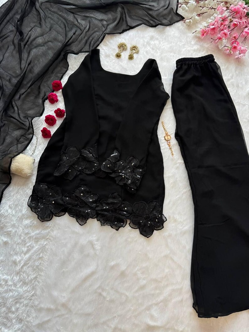 May include: A black two-piece outfit with a long-sleeved top and wide-leg pants. The top features floral lace detailing with sequins at the hem. A sheer black scarf and matching pants complete the ensemble. Gold earrings and a bracelet are also visible.