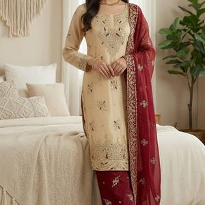 Puede incluir: Un conjunto salwar kameez beige bordado con un dupatta granate. La t&uacute;nica y el pantal&oacute;n de manga larga est&aacute;n adornados con intrincados bordados dorados. La bufanda granate transparente tiene un borde dorado. El atuendo es adecuado para ocasiones especiales.