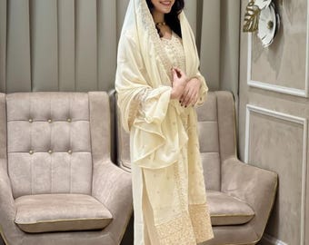 May include: Cream-colored embroidered ensemble featuring a tunic, pants, and a head covering. The outfit has intricate gold embroidery along the edges and sleeves. The person is wearing gold-colored sandals. The background includes two beige armchairs and a wall with floral decorations.