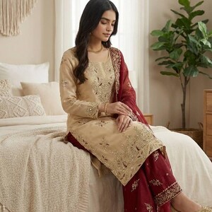 Puede incluir: Un conjunto salwar kameez beige bordado con pantalones palazzo burdeos y una dupatta a juego. El atuendo presenta intrincados bordados dorados y se usa con bailarinas doradas. El conjunto es adecuado para ocasiones festivas.