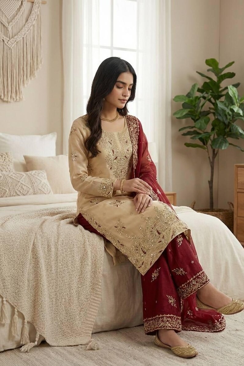 Puede incluir: Un conjunto salwar kameez beige bordado con pantalones palazzo burdeos y una dupatta a juego. El atuendo presenta intrincados bordados dorados y se usa con bailarinas doradas. El conjunto es adecuado para ocasiones festivas.