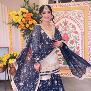 May include: A woman wearing a traditional South Asian outfit. The ensemble includes a white and gold top, a matching skirt, and a dark blue shawl with a floral pattern. The outfit is complemented by a headpiece.