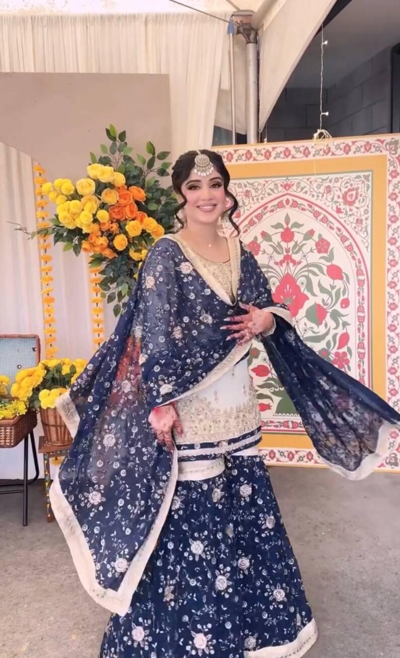 May include: A woman wearing a traditional South Asian outfit. The ensemble includes a white and gold top, a matching skirt, and a dark blue shawl with a floral pattern. The outfit is complemented by a headpiece.