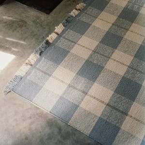 K&ouml;nnte beinhalten: Ein blau-wei&szlig; karierter Teppich mit Fransen. Der Teppich hat ein gro&szlig;es Karomuster in Blau- und Off-White-T&ouml;nen. Der Teppich besteht aus gewebtem Material und liegt auf einer Betonoberfl&auml;che.