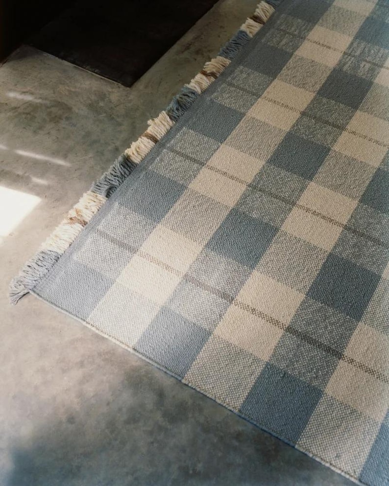 K&ouml;nnte beinhalten: Ein blau-wei&szlig; karierter Teppich mit Fransen. Der Teppich hat ein gro&szlig;es Karomuster in Blau- und Off-White-T&ouml;nen. Der Teppich besteht aus gewebtem Material und liegt auf einer Betonoberfl&auml;che.