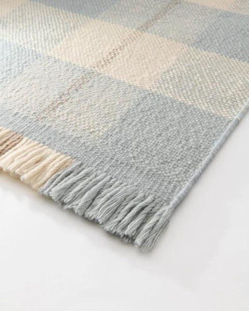 K&ouml;nnte beinhalten: Nahaufnahme einer gewebten Decke mit einem karierten Muster in den Farbt&ouml;nen Hellblau, Creme und Hellbraun. Die Decke hat einen Fransenrand in Hellblau. Die Textur der Decke wirkt weich und gem&uuml;tlich.