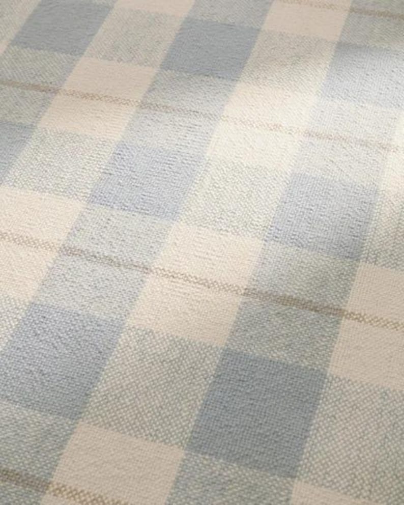 K&ouml;nnte beinhalten: Nahaufnahme eines gewebten Stoffes mit gro&szlig;em Karomuster. Das Design zeigt abwechselnde Quadrate in Hellblau, Off-White und einem dezenten Braunton. Der Stoff hat ein strukturiertes Aussehen, was auf ein weiches, bequemes Material hindeutet.