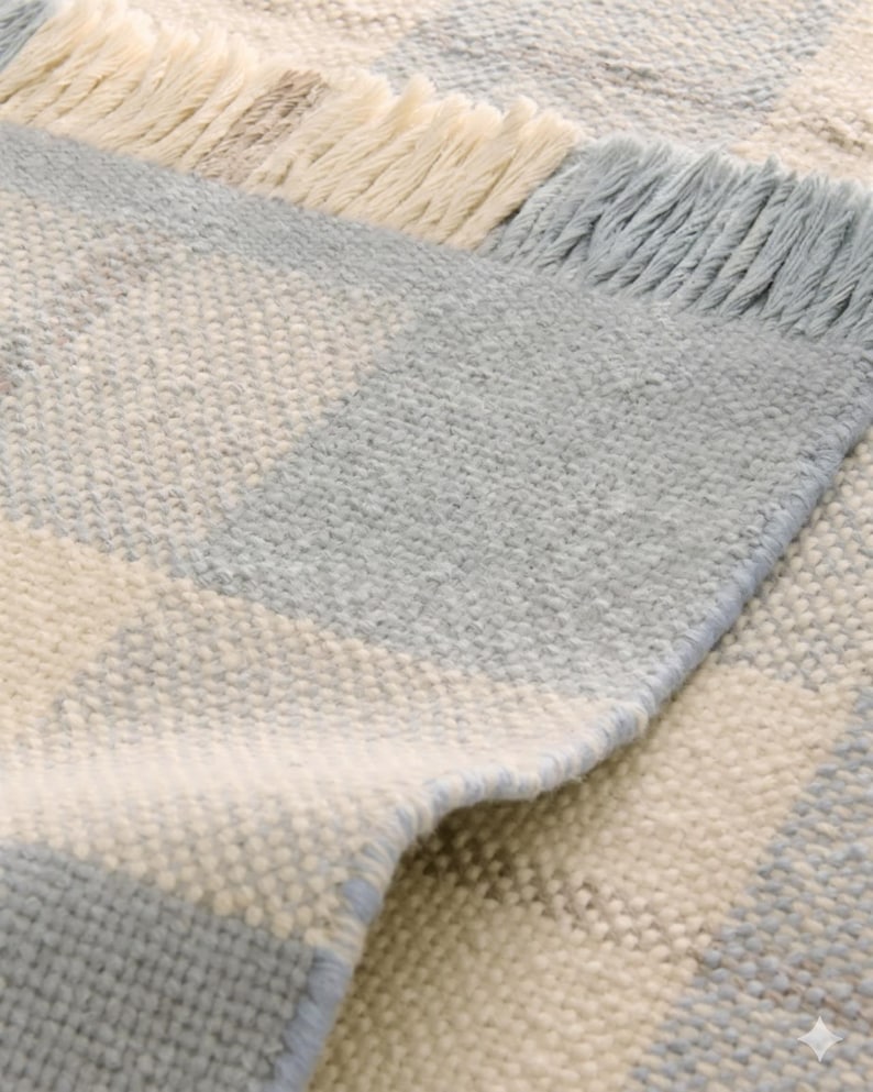 K&ouml;nnte beinhalten: Nahaufnahme eines gewebten Teppichs mit geometrischem Muster. Der Teppich weist abwechselnde Streifen in Hellblau, Off-White und einem Hauch von Braun auf. Die Kanten sind mit einem Fransenbesatz in passenden Farben versehen, der Textur und Detail verleiht.