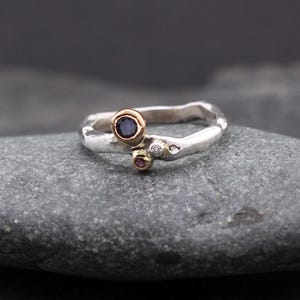 Peut inclure: Bague en argent avec une bande texturée, ornée d'une pierre précieuse bleu foncé sertie dans une lunette dorée, d'une petite pierre précieuse rouge et d'une minuscule pierre précieuse blanche. La bague est présentée sur une surface en pierre grise.