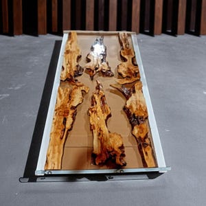 May include: A rectangular wooden table with a clear resin top. The table features six pieces of natural wood with a light brown color and dark brown accents. The table is set on a gray surface.
