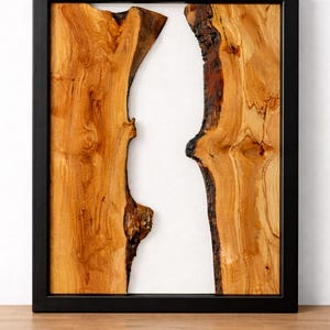 May include: A framed artwork featuring two pieces of natural wood with a light brown color and dark brown edges, set against a white background. The wood grain is visible. The frame is black.