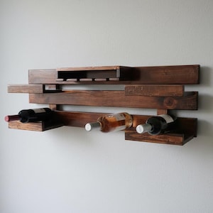 Può includere: Un portabottiglie da vino in legno tinto scuro montato a parete. Il portabottiglie può contenere tre bottiglie di vino e ha una sezione per appendere i calici. Il design è moderno e geometrico.