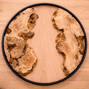 May include: A circular wooden serving tray with a black rim. The tray has a light wood centre with two organic-shaped pieces of textured, light brown wood. The tray is set on a light wood surface.