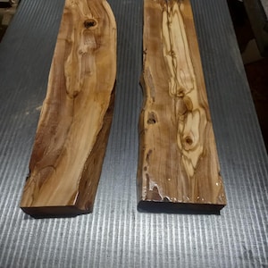 May include: Two rectangular pieces of olive wood with natural live edges. The wood displays a polished, glossy finish highlighting the intricate brown and cream grain patterns, knots, and organic swirls characteristic of the olive tree species.