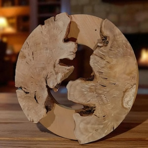 Könnte beinhalten: Runde Wanddekoration aus hellem Holz mit natürlicher Maserung. Das Design weist eine zentrale Öffnung und einen rauen, strukturierten Rand auf. Das Stück wird auf einer Holzoberfläche präsentiert.