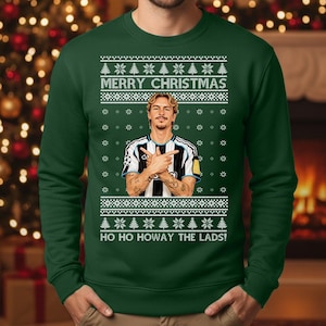 Puede incluir: Jersey navideño verde oscuro con un diseño festivo. El jersey presenta el texto "MERRY CHRISTMAS" y "HO HO HOWAY THE LADS!". También incluye un gráfico de una persona con una camiseta a rayas blancas y negras. El jersey es de cuello redondo.