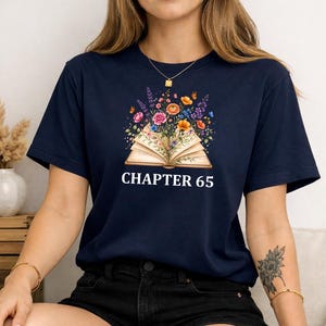 May include: A navy blue short-sleeve t-shirt featuring a graphic of an open book with colorful flowers and butterflies emerging from the pages. The text "CHAPTER 65" is printed in white capital letters directly below the illustrated book graphic.