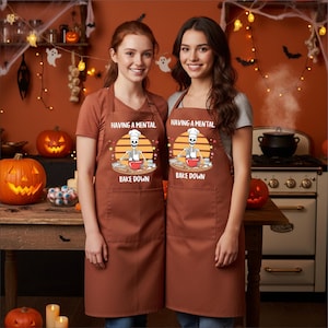 Puede incluir: Dos mujeres con delantales marrones con el texto "HAVING A MENTAL BAKE DOWN" y un gráfico de un chef esqueleto. Los delantales tienen grandes bolsillos delanteros. El fondo incluye decoraciones de Halloween como calabazas y telarañas.