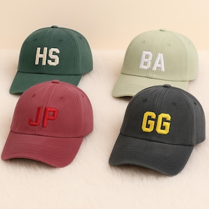 Könnte beinhalten: Vier Baseballkappen in verschiedenen Farben: Dunkelgrün, Hellgrün, Burgunderrot und Anthrazit. Jede Kappe hat gestickte Blockbuchstaben: "HS", "BA", "JP" und "GG". Die Kappen haben gebogene Krempen und verstellbare Riemen.