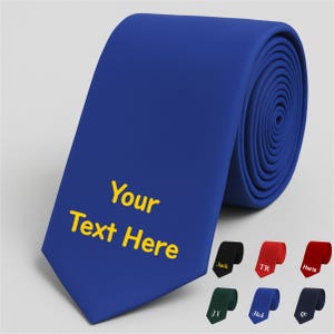 Könnte beinhalten: Eine gerollte, königsblaue Krawatte mit dem Text "Your Text Here" in Gelb. Zusätzliche Krawatten in Schwarz, Rot, Grün und Marineblau werden darunter angezeigt. Die Krawatten sind ein klassisches Accessoire.