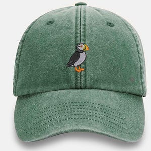 Puede incluir: Gorra de béisbol verde con un diseño bordado de frailecillo. El frailecillo tiene plumas negras, blancas y grises, un pico y patas naranjas. La gorra tiene una visera curva y una correa ajustable.