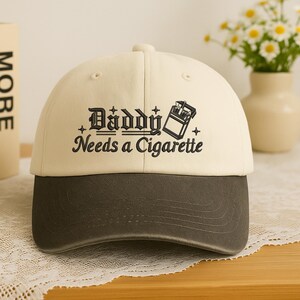 May include: A baseball cap with a cream-colored crown and a dark gray brim. The cap features the text "Daddy Needs a Cigarette" in black, along with a graphic of a cigarette pack.