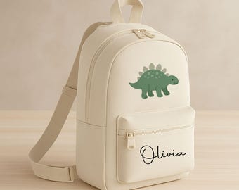 May include: Cream-colored backpack featuring a green dinosaur illustration and the name "Olivia" in black script. The backpack has a front zippered pocket, a top handle, and adjustable shoulder straps. The zippers are a light beige color.
