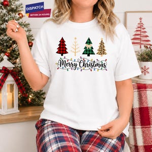 Può includere: Una t-shirt bianca a maniche corte con una grafica di quattro alberi di Natale stilizzati a quadri rossi, contorno dorato, quadri verdi con un fiocco e stampa leopardata. Sotto gli alberi, la scritta Merry Christmas è in corsivo nero.