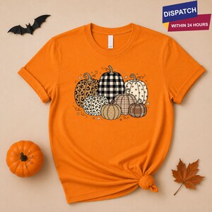 May include: An orange short-sleeve t-shirt featuring a graphic print of seven pumpkins with various patterns including leopard print, black and white buffalo plaid, polka dots, and solid brown, arranged in a cluster with small star accents.
