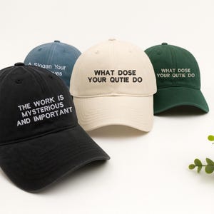 Op de afbeelding: Vier baseballpetten in zwart, crème, blauwgroen en donkergroen. Elke pet heeft witte tekst. Op de zwarte pet staat "THE WORK IS MYSTERIOUS AND IMPORTANT". Op de crème en groene petten staat "WHAT DOSE YOUR QUTIE DO". Op de blauwgroene pet staat "A Slogan Your ves."