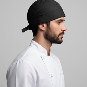Puede incluir: Un gorro de cocinero negro con una cinta en la parte trasera. El gorro se lleva con una chaqueta de cocinero blanca con cuello mandar&iacute;n y cierre de botones. El fondo es gris claro liso.
