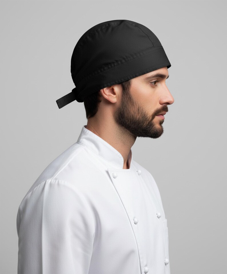 Puede incluir: Un gorro de cocinero negro con una cinta en la parte trasera. El gorro se lleva con una chaqueta de cocinero blanca con cuello mandar&iacute;n y cierre de botones. El fondo es gris claro liso.