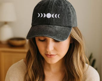 May include: A dark gray baseball cap with a curved brim. The cap features a white embroidered design of the phases of the moon across the front. The hat is made of a soft, worn material.