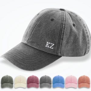 Puede incluir: Gorra de béisbol gris con las iniciales blancas "KZ". La gorra tiene una visera curva y un cierre de correa de tela. Debajo, seis gorras en diferentes colores: verde, crema, burdeos, gris, azul, rosa y óxido.